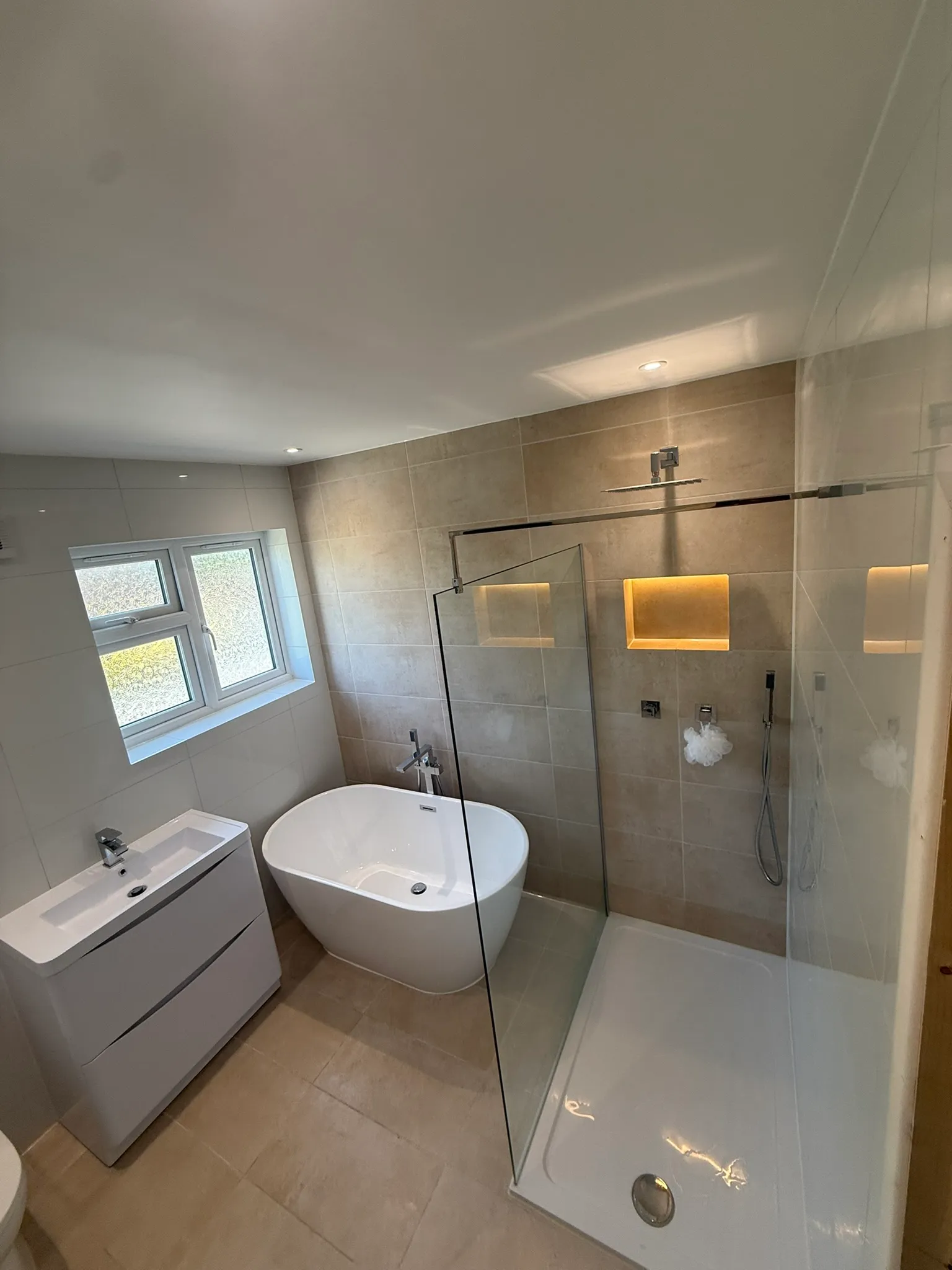 Fitted bathroom with a freestanding oval bath, floor-standing chrome bath tap, wall-hung vanity unit and a glass-screened walk-in shower with lit tiled niches